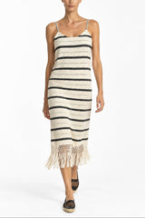 A woman modeling a sleeveless knit midi dress with horizontal black stripes on an off-white base, featuring thin straps and a fringed hem, set against a plain white studio background.