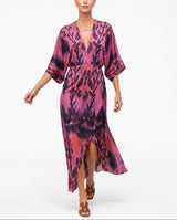 A woman modeling a long-sleeved, wrap-style midi dress with a pink, purple, and black abstract floral print, set against a plain white studio background.