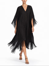 A woman modeling a black fringed kaftan dress with a V-neckline and wide sleeves, styled with black strappy high-heeled sandals against a plain white studio background.