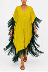 A woman modeling a bright yellow kaftan-style poncho dress with dark green fringe trim along the sleeves and hem, set against a plain light gray studio background.