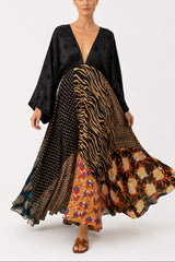 A model wearing a long, flowing maxi dress with a mixed print design, featuring a black velvet long-sleeve top and a skirt with zebra, polka dot, floral, and orange patterns, set against a plain light gray studio background.