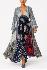 A woman modeling a long, flowing maxi dress with mixed prints, including navy blue with white palm leaves, gray and white geometric patterns, and a vibrant magenta floral section, set against a plain light gray studio background.