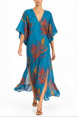 A woman modeling a flowing teal blue kaftan dress with a vibrant orange and yellow floral pattern, set against a plain white studio background.