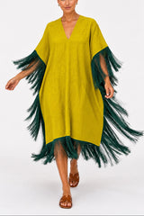 A woman modeling a loose, V-neck kaftan dress in mustard yellow with dark green feathered fringe along the sleeves and hem, paired with brown leather slide sandals, against a plain white studio background.